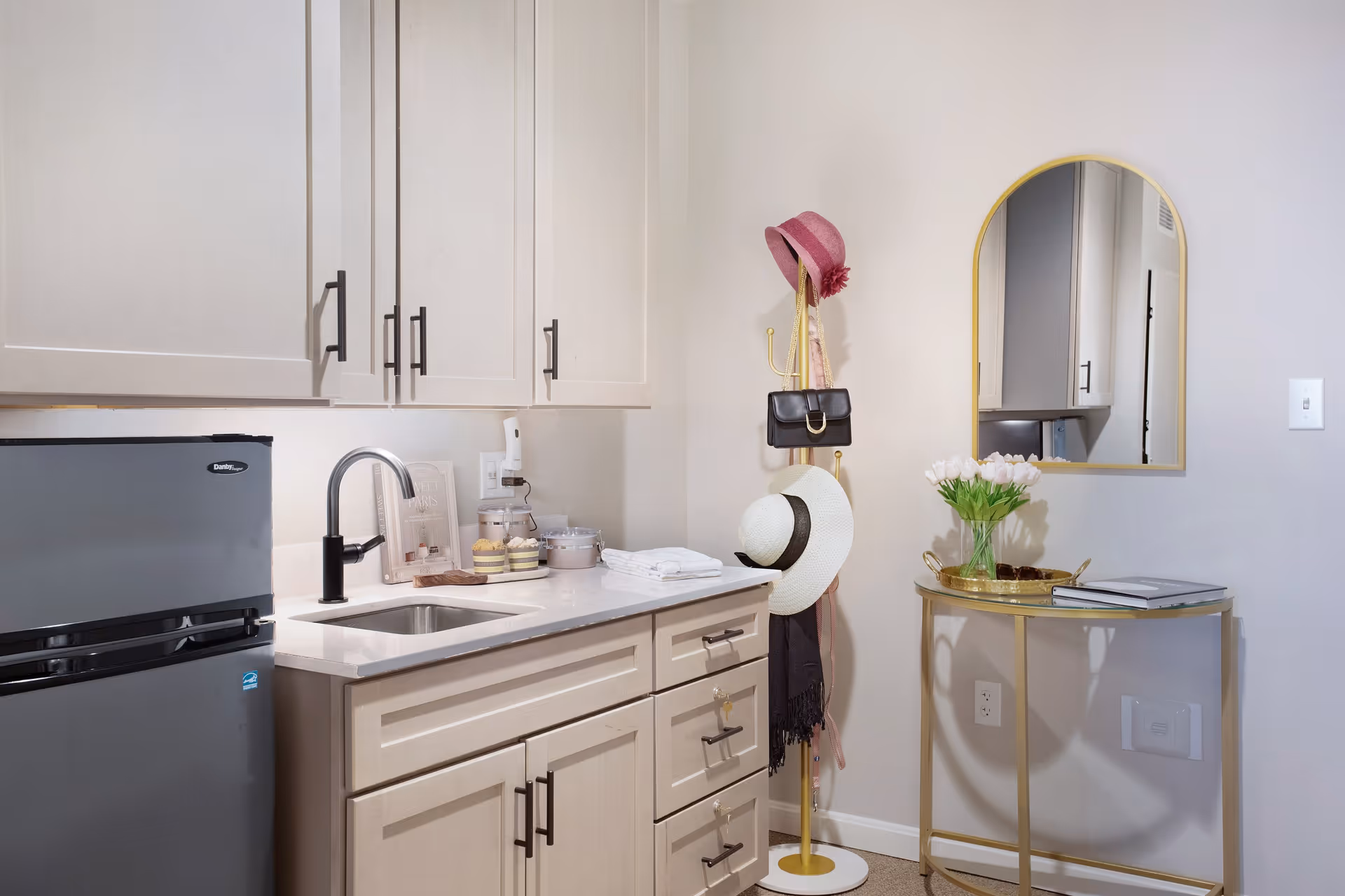A small kitchenette area with light-colored cabinets, a stainless steel mini refrigerator, a sink with a modern faucet, and countertop items including jars and a book. To the right, there is a gold coat rack holding hats, a purse, and a scarf. Next to it is a gold-framed arched mirror above a small gold table with a vase of white tulips and an open book.