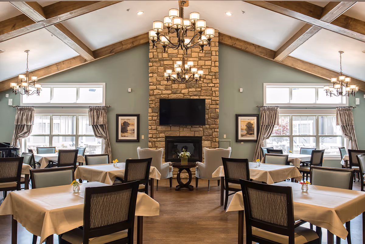 A spacious dining room with multiple tables covered in white tablecloths, each surrounded by chairs. The room features a high ceiling with wooden beams and three elegant chandeliers. A stone fireplace with a mounted flat-screen TV is centered on the far wall, flanked by two framed pictures and large windows with curtains allowing natural light to enter.