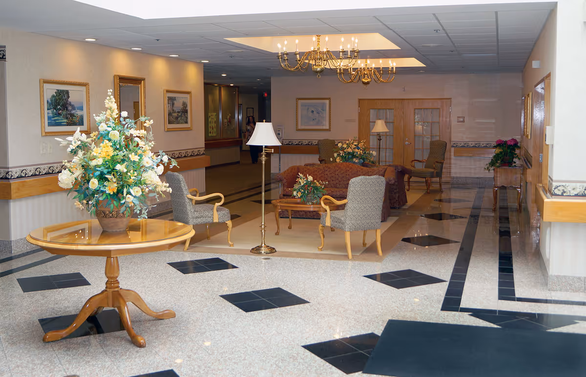 A well-lit interior common area with a polished tile floor featuring black diamond patterns. In the foreground, there is a round wooden table with a large floral arrangement. The background shows a seating area with upholstered chairs, a sofa, a coffee table with flowers, a floor lamp, and a chandelier hanging from the ceiling. The walls are decorated with framed paintings and mirrors.