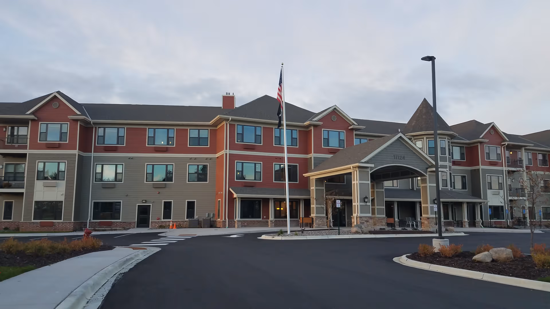 Exterior view of a three-story senior living facility building with red and gray siding, multiple windows, a covered entrance with the number 11124, an American flag on a flagpole, and a paved driveway with landscaping.
