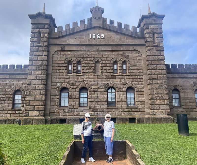 Two elderly women standing in front of a historic stone building with the year 1862 displayed at the top. The building has arched windows and a castle-like design with battlements. The women are standing on a grassy area near an old cannon.