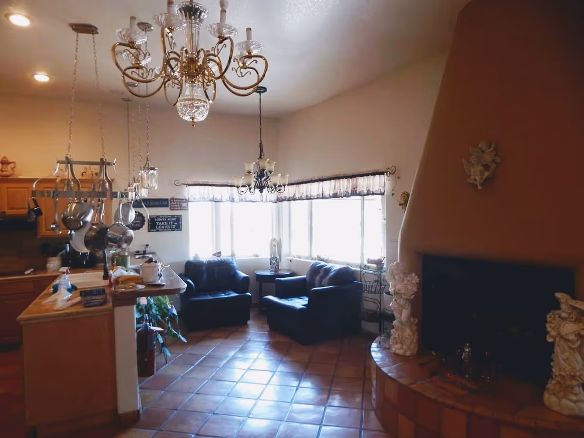 Interior view of a cozy living space with a kitchen area on the left, featuring hanging pots and pans above a counter. Two black armchairs and a small round table are positioned near a large window with sheer curtains. A decorative fireplace with angel figurines is on the right side. The room has tiled flooring and two ornate chandeliers hanging from the ceiling.