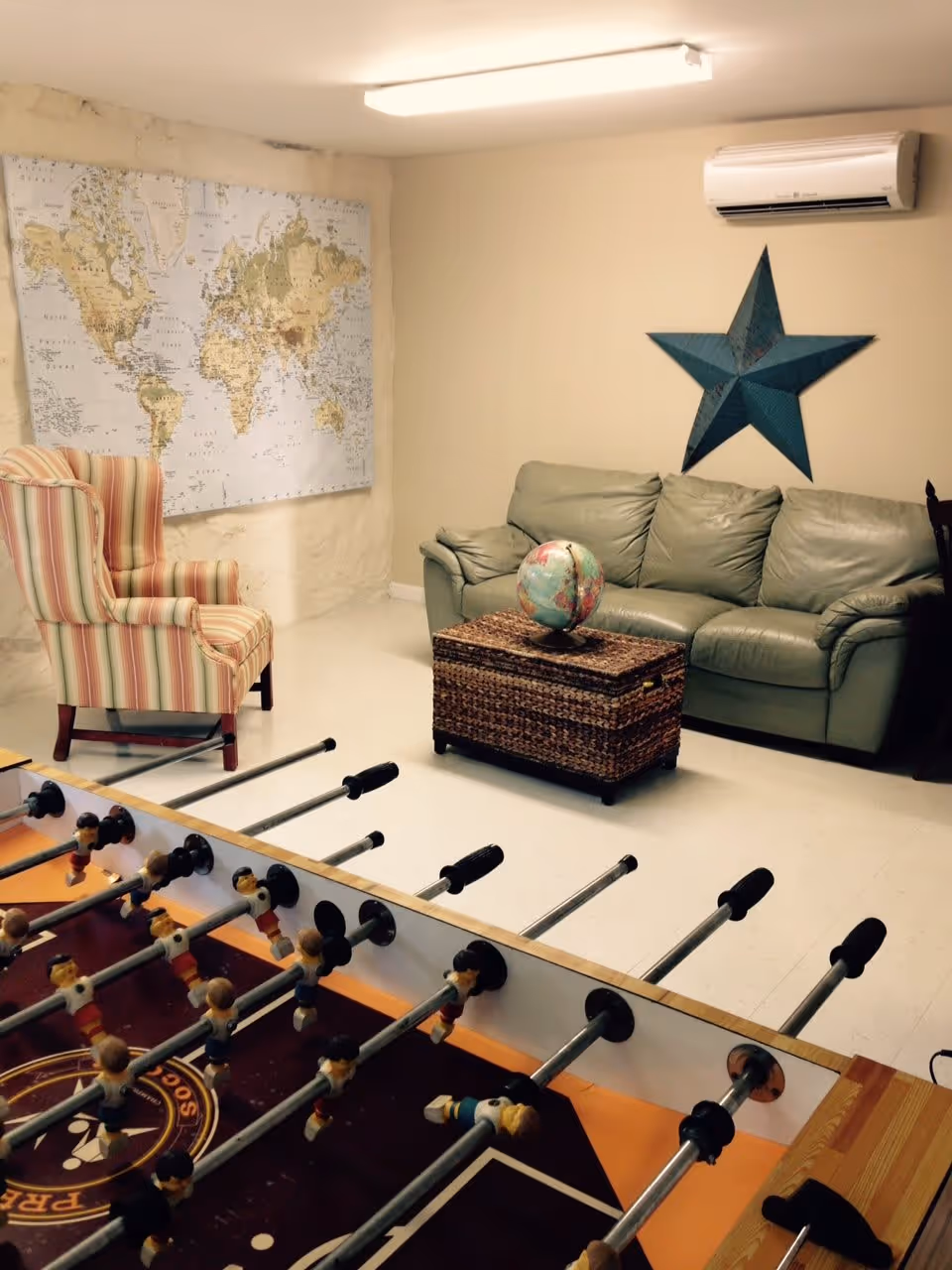 A cozy recreational room with a foosball table in the foreground, a striped armchair, a green leather couch, a wicker coffee table with a globe on top, a large world map on the wall, and a blue star decoration mounted on the wall beneath an air conditioning unit.