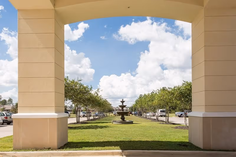View through a large archway showing a green lawn with a multi-tiered water fountain in the center, flanked by rows of small trees and parked cars on either side under a partly cloudy blue sky.