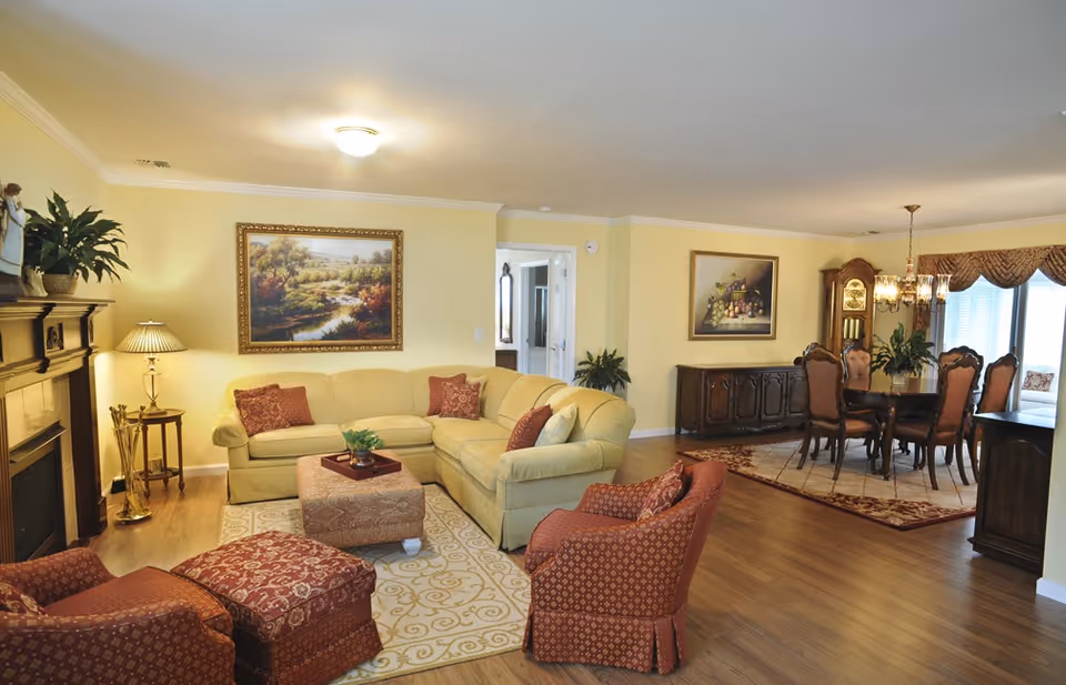Open-plan living and dining area with yellow walls, a beige sectional sofa, red patterned armchairs, fireplace, artwork, and a wooden dining table.