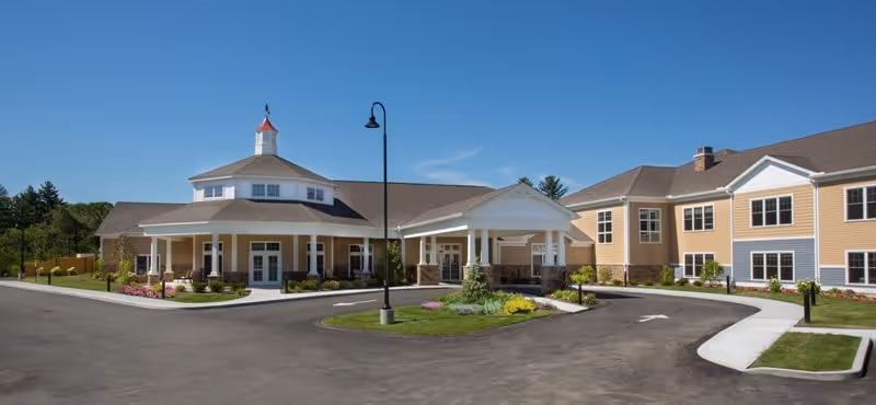 Front entrance of a large yellow senior living building with a covered porte-cochère, circular drive and landscaped grounds under a clear blue sky.