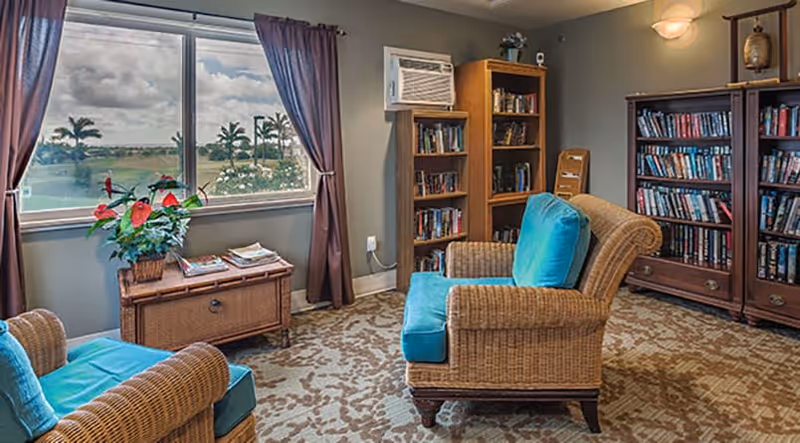 A cozy reading room with two wicker armchairs featuring teal cushions, a wicker side table with a potted plant and magazines, two wooden bookshelves filled with books, a window with brown curtains showing a view of palm trees and a cloudy sky, and a wall-mounted air conditioning unit.