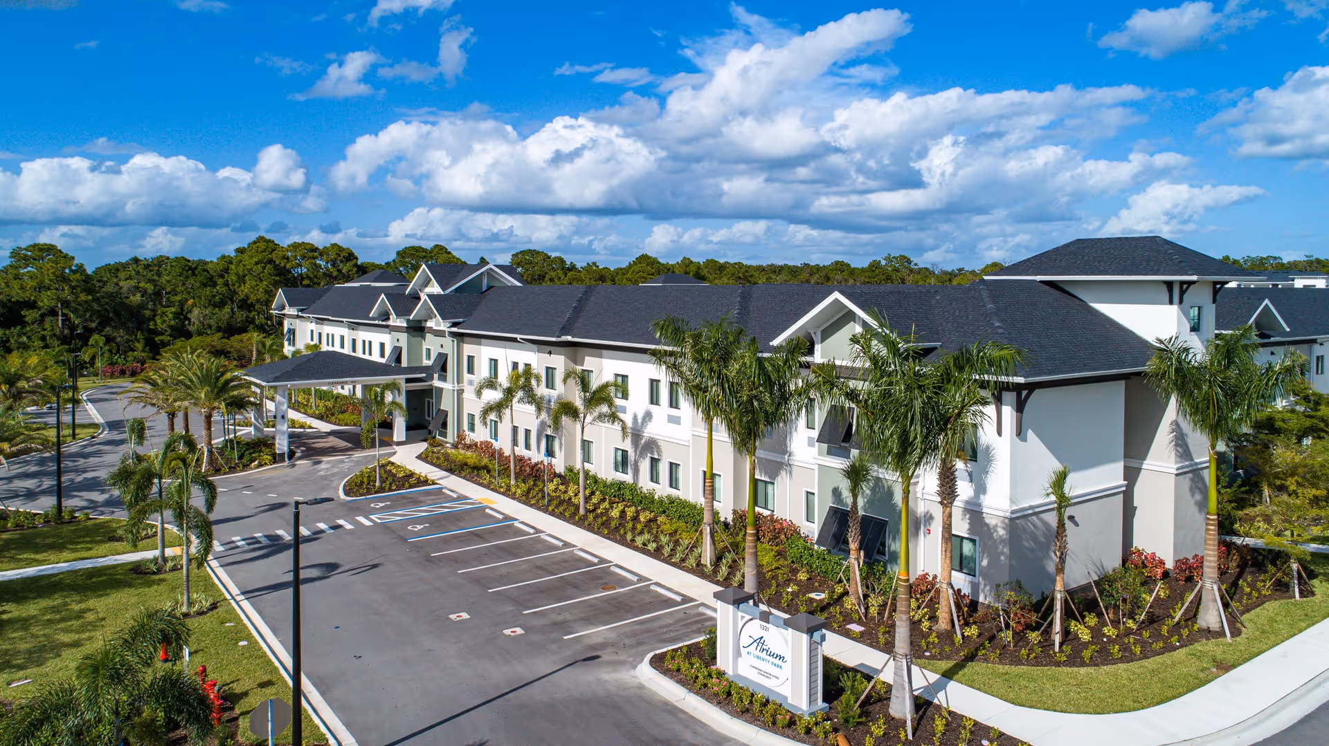 Exterior view of a modern two-story senior living facility named Atrium at Liberty Park, surrounded by palm trees and landscaped greenery under a partly cloudy blue sky. The building has a covered entrance and an adjacent parking lot with marked spaces.