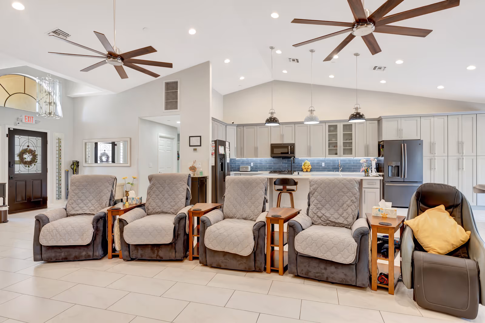 A bright and spacious assisted living common area featuring four cushioned recliners with quilted covers and wooden side tables, a black leather chair with a yellow pillow, a modern kitchen with gray cabinetry, blue tile backsplash, and stainless steel appliances in the background, and two large ceiling fans overhead.