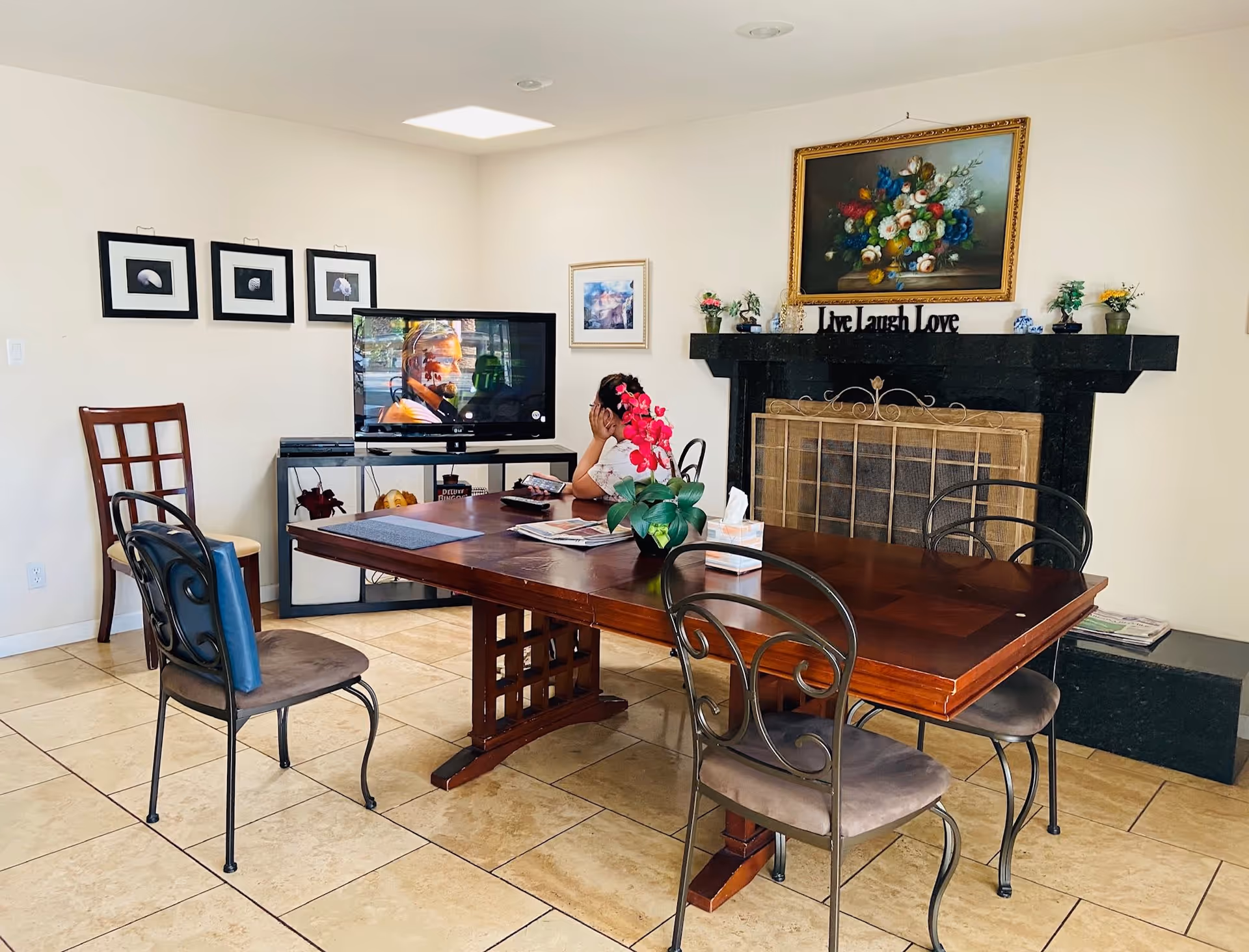 A cozy living room with a wooden dining table surrounded by four chairs. A young girl is sitting at the table watching a television placed on a stand against the wall. The room features a black fireplace with a decorative screen and a floral painting above it. The walls are adorned with framed pictures, and there is a pink orchid plant on the table.