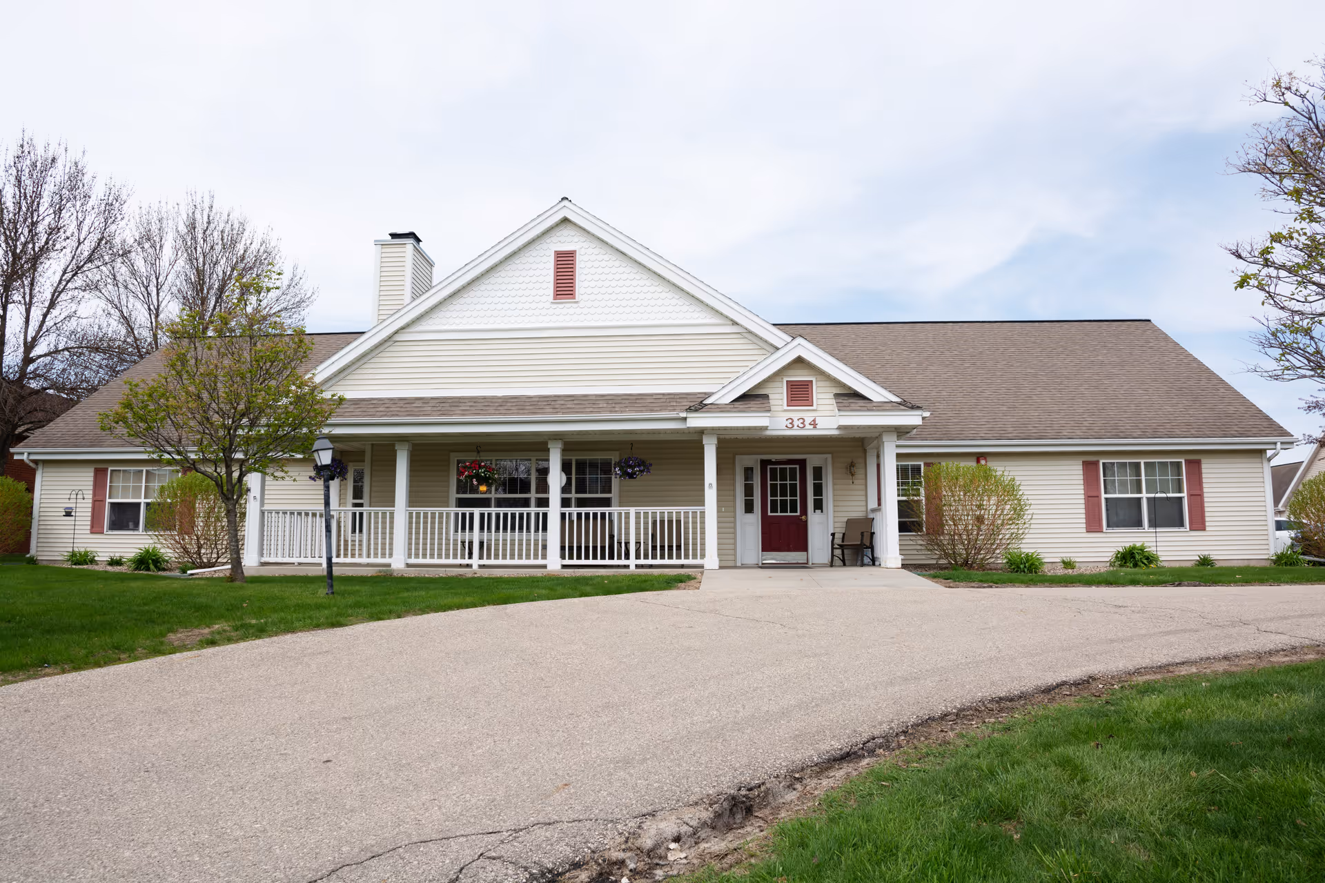 Front exterior view of a single-story building with beige siding, a brown roof, and a covered porch with white railings. The building has red shutters and a red door with the number 334 above it. There are small trees and bushes on either side of the entrance, and a paved driveway leading up to the porch.