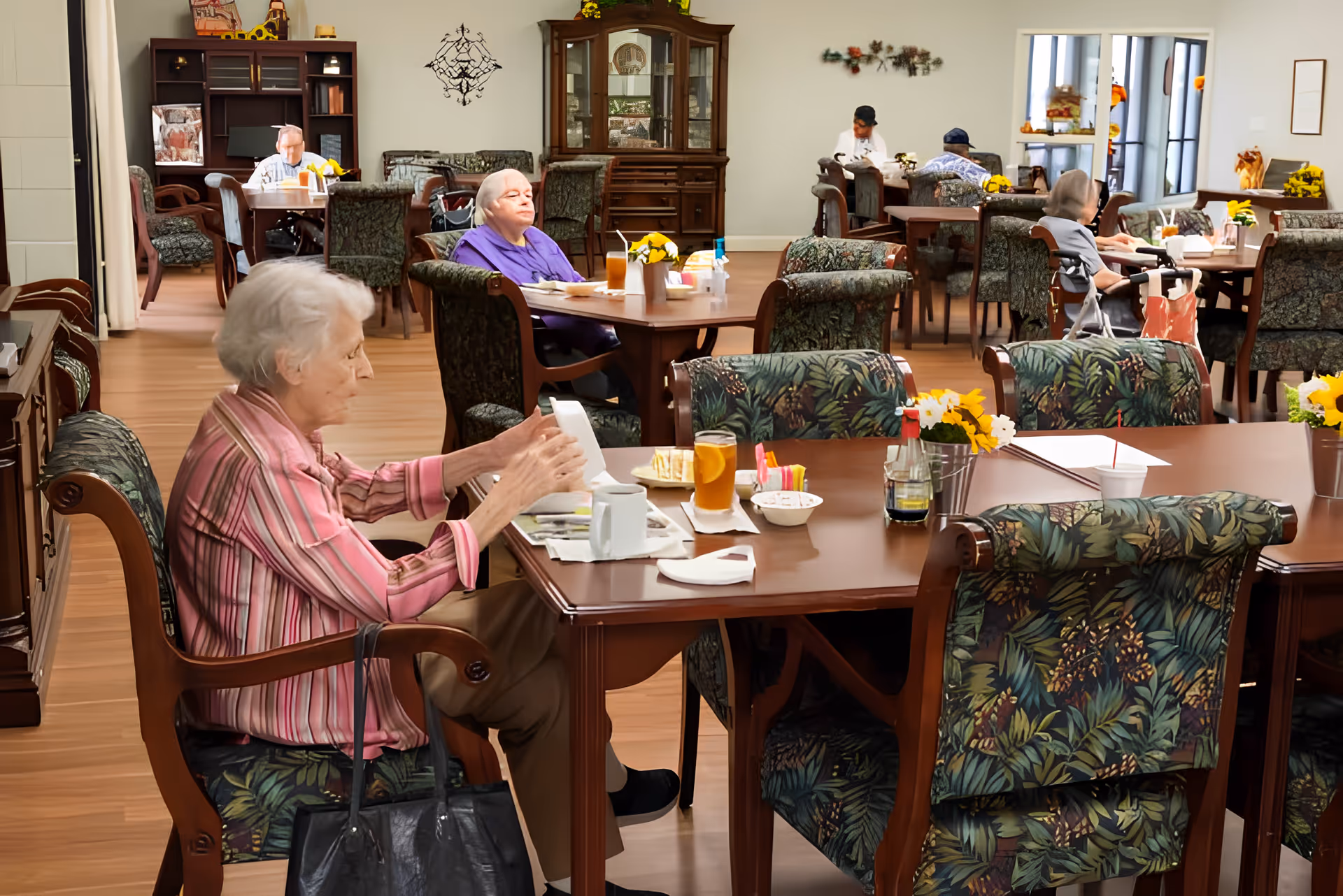 A dining room in a senior living facility with elderly residents seated at tables. The room has wooden floors, floral-patterned chairs, and tables set with drinks, flowers, and condiments. In the background, there is a wooden cabinet and wall decorations.