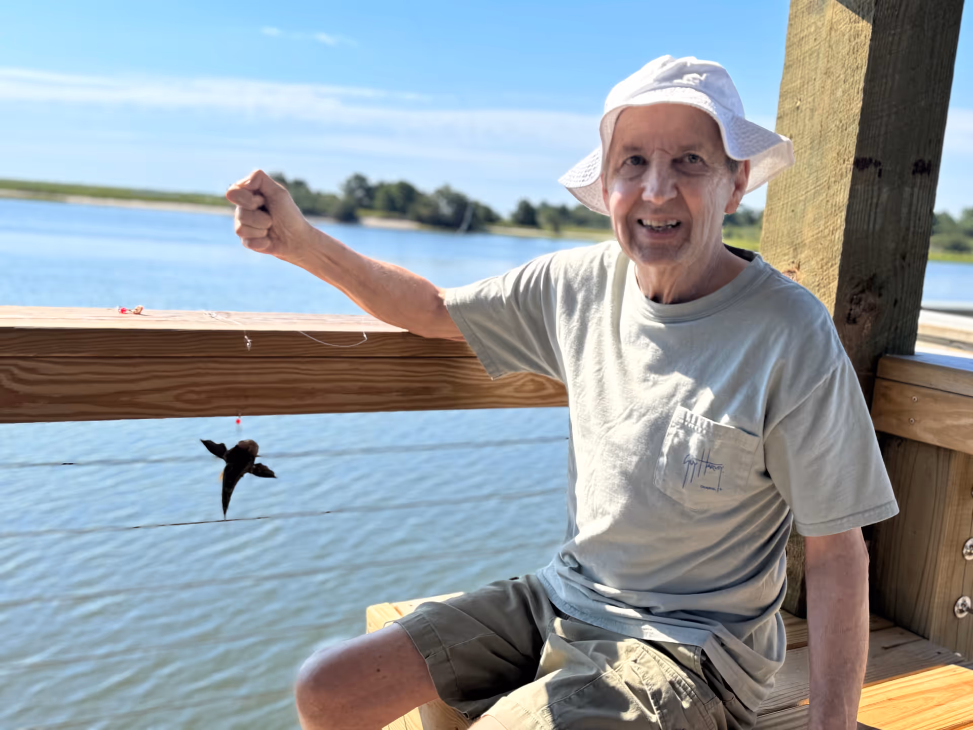 An elderly man wearing a white bucket hat and light-colored t-shirt sits on a wooden bench on a dock by the water, holding up a fishing line with a small fish caught on the hook. The background shows a calm body of water and a clear blue sky with some trees in the distance.