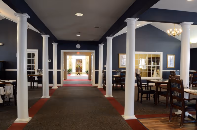 Interior view of a dining area with multiple wooden tables and chairs arranged neatly. The room features white columns, dark blue walls, and a carpeted floor with red borders. A chandelier hangs from the ceiling on the right side, and a hallway with a floral arrangement at the end is visible in the background.