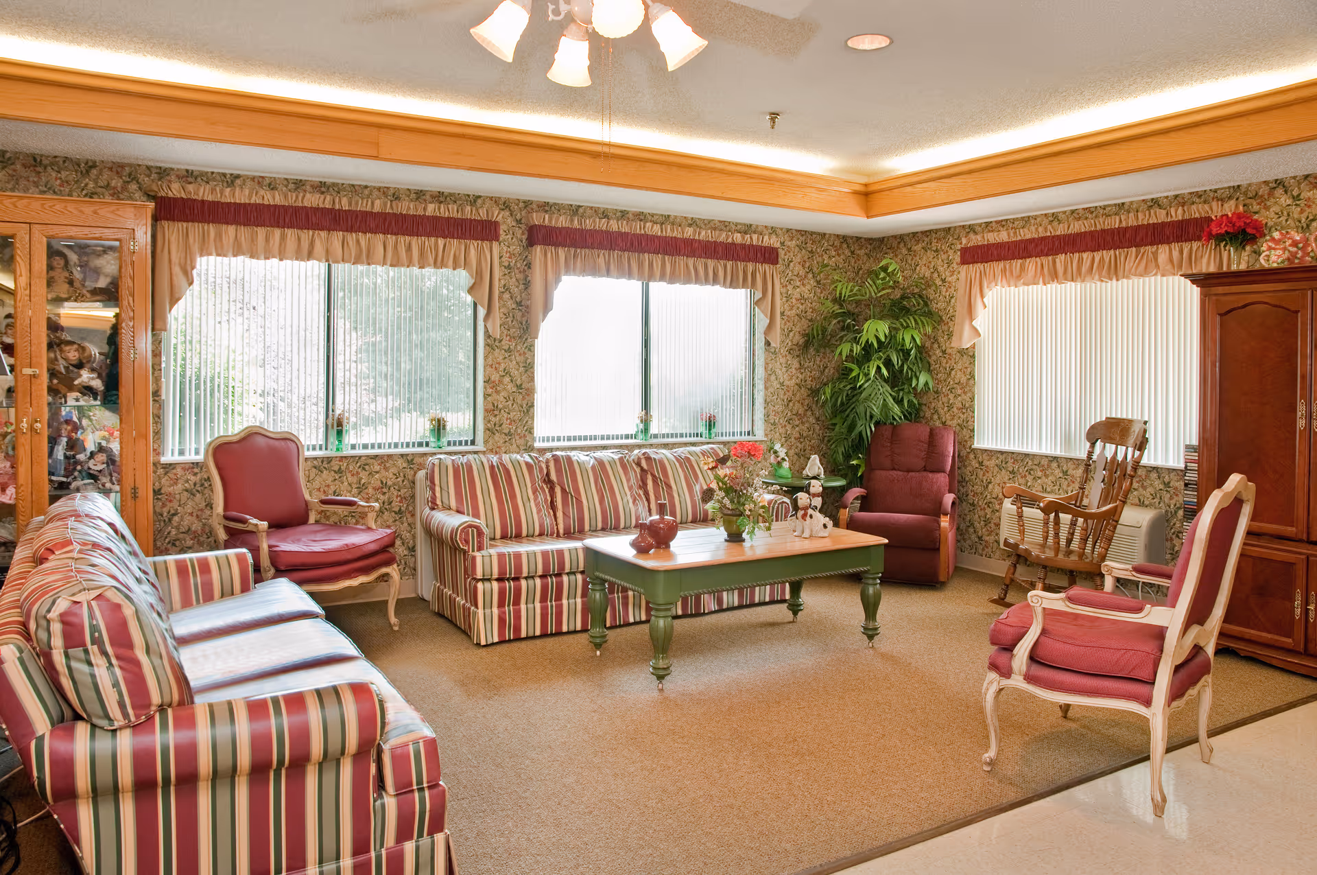 Well-lit communal living room with striped sofas and armchairs arranged around a green coffee table, with large windows and decorative plants.