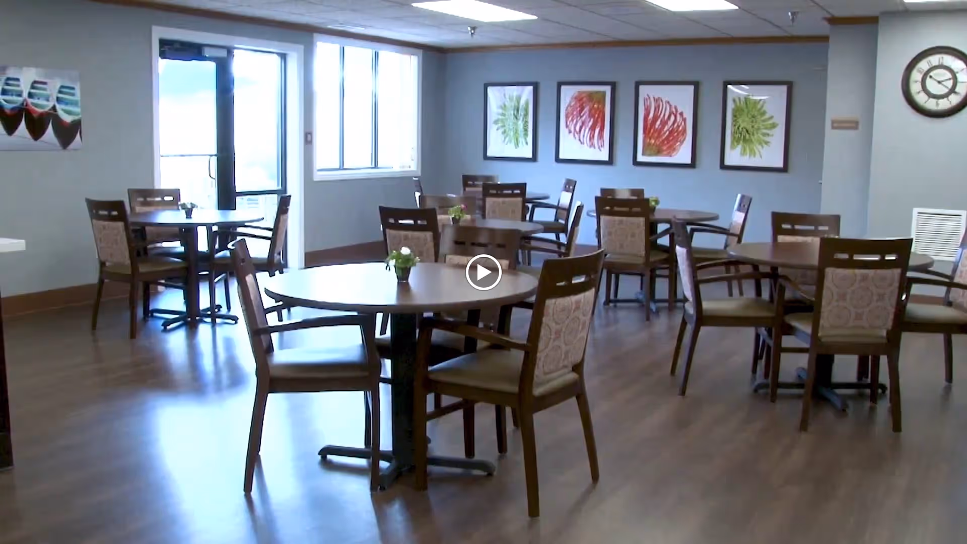 A dining room with multiple round wooden tables and chairs arranged neatly. Each table has a small flower centerpiece. The room has large windows letting in natural light, framed botanical artwork on the walls, and a clock mounted on the right wall.