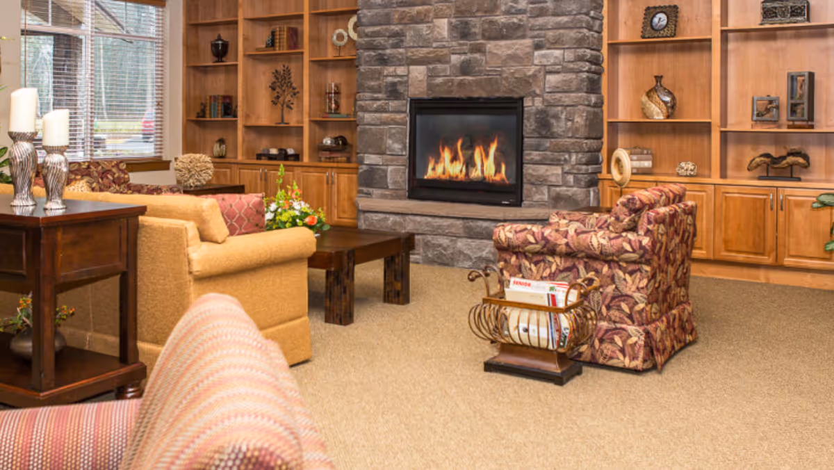 A cozy living room with a stone fireplace in the center, surrounded by wooden built-in shelves and cabinets. The room features comfortable upholstered armchairs and a sofa in warm tones, a wooden coffee table, decorative items on the shelves, and a magazine rack near one of the chairs. Large windows with blinds allow natural light to enter the space.