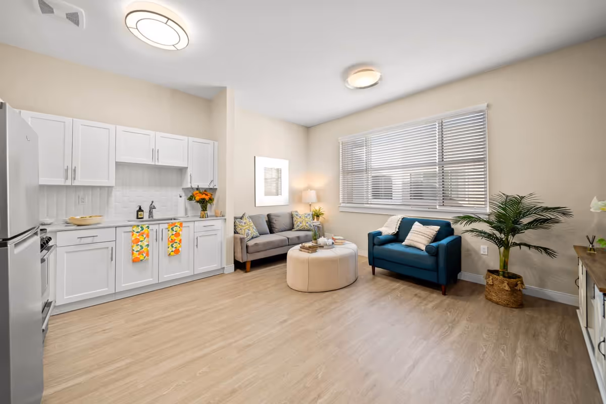 A bright and modern living space featuring a small kitchen area with white cabinets, a refrigerator, and a stove on the left. The room has light wood flooring and beige walls. There is a gray sofa with patterned pillows and a blue armchair with a pillow near a large window with blinds. A round beige ottoman is placed in the center with books and a tea set on top. A potted plant is positioned near the armchair, and a lamp with a white shade is on a side table next to the sofa.