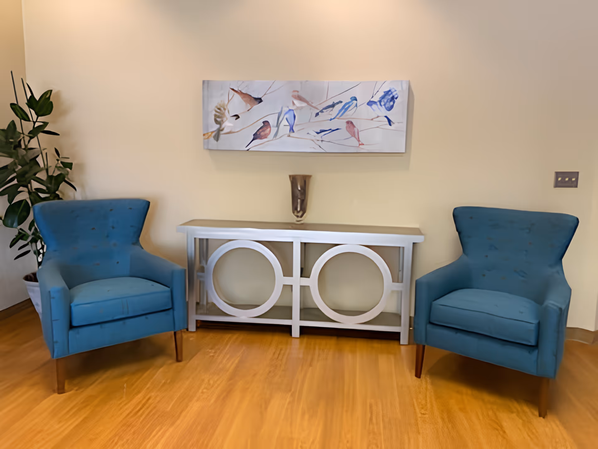Two blue upholstered armchairs positioned on either side of a white console table with circular design elements. A decorative vase sits on the table, and a painting of various birds on branches hangs on the beige wall above. A green potted plant is visible to the left of the left chair, and the floor is wooden.