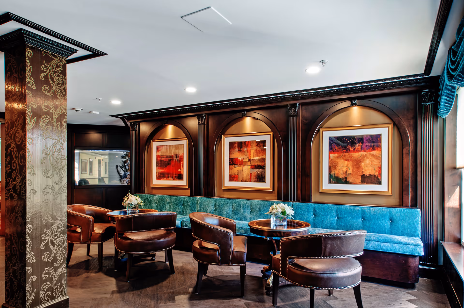A cozy seating area with a turquoise upholstered bench along the wall and four brown leather chairs around two small round wooden tables, each with a small flower arrangement. The wall behind the bench features three framed abstract paintings with warm colors, set in arched wooden panels with decorative molding. The room has a polished wooden floor and a column with ornate wallpaper pattern on the left side.