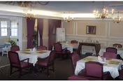 A dining room with multiple round tables covered with white tablecloths and set with placemats. Each table is surrounded by purple upholstered chairs. The room has beige walls with white trim, chandeliers hanging from the ceiling, and large windows with purple curtains. There is a sideboard against one wall with a mirror above it and a flip chart stand nearby.