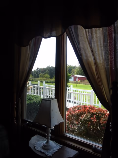 Interior view through a curtained window with a small lamp on a table and a white porch railing, shrubs, lawn, and a red outbuilding outside.