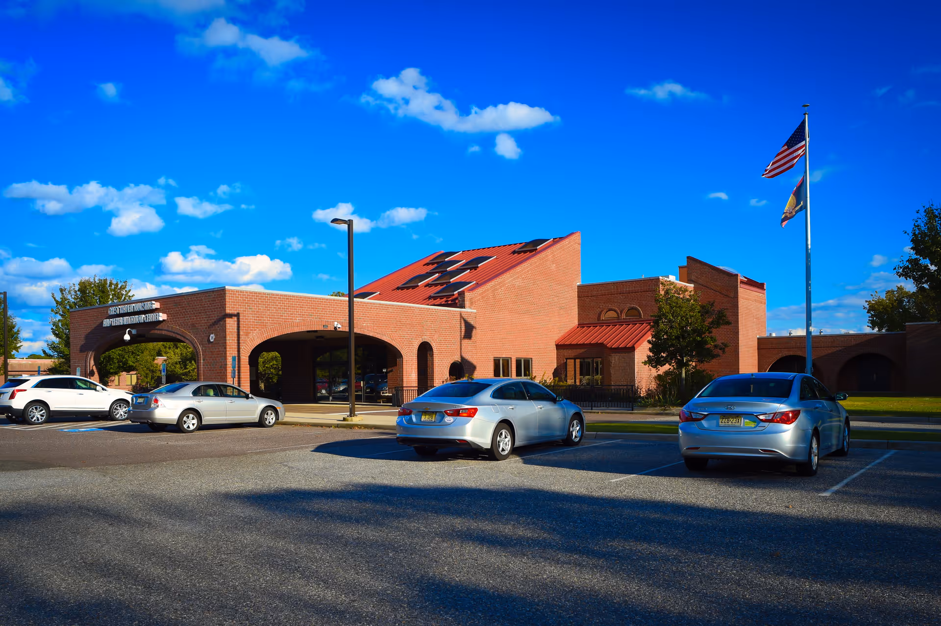 Exterior view of Crest Haven Nursing & Rehabilitation Center, a brick building with a red roof and solar panels. Several cars are parked in front, and two flagpoles display the American flag and another flag. The sky is clear with a few clouds.
