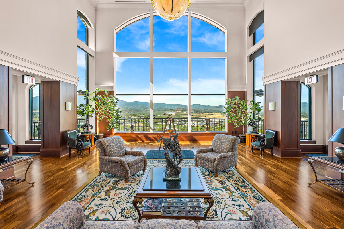 A spacious and elegant living room with large floor-to-ceiling windows offering a panoramic view of mountains and a valley. The room features wooden floors, patterned armchairs, a glass coffee table with a decorative sculpture, potted plants, and warm wood paneling on the walls. Two exit signs are visible above doorways on either side of the room.