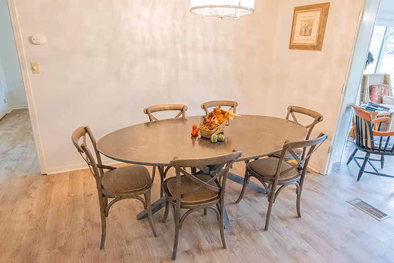 A dining area with a large oval wooden table surrounded by six wooden chairs with woven seats. A decorative centerpiece with autumn-themed items is placed on the table. The room has light-colored walls and wood flooring. A framed picture hangs on the wall, and an adjacent room with additional seating is visible through an open doorway.