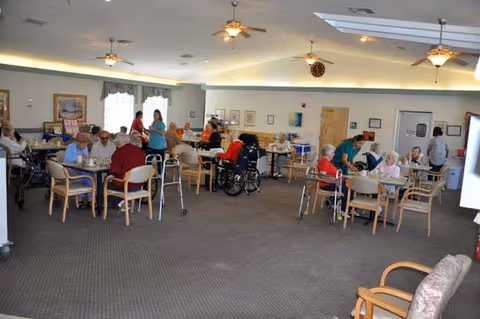 A spacious dining room in an assisted living facility with several elderly residents seated at tables, some using walkers and wheelchairs. Staff members are assisting and interacting with the residents. The room has ceiling fans, framed pictures on the walls, and large windows letting in natural light.