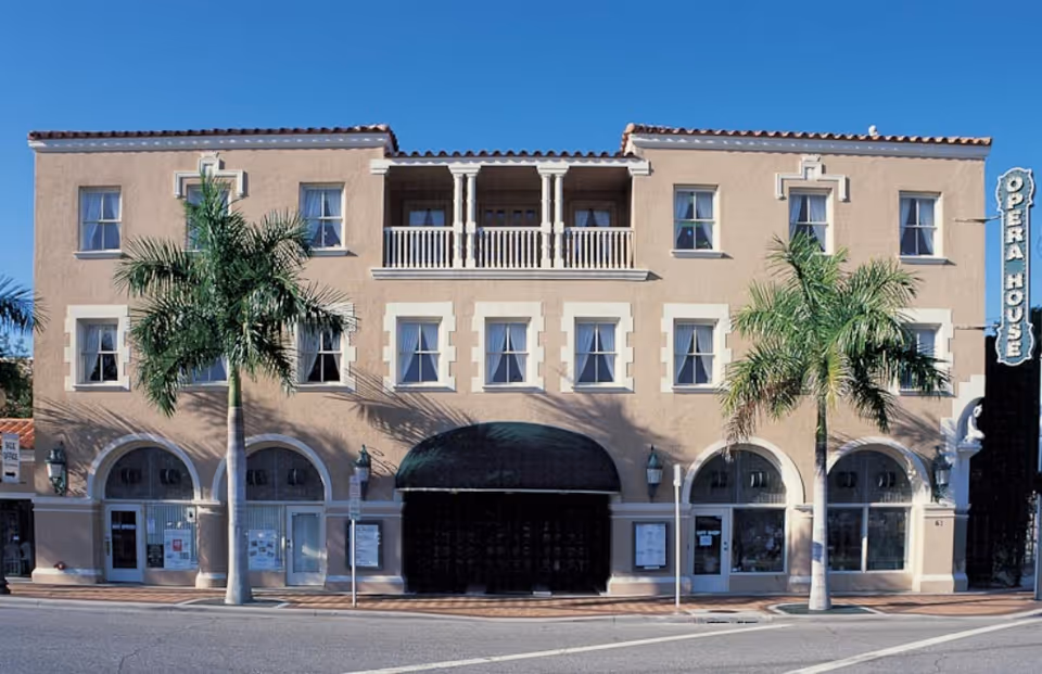Three-story Mediterranean-style building with arched ground-floor storefronts, two palm trees, and a vertical 'OPERA HOUSE' sign.