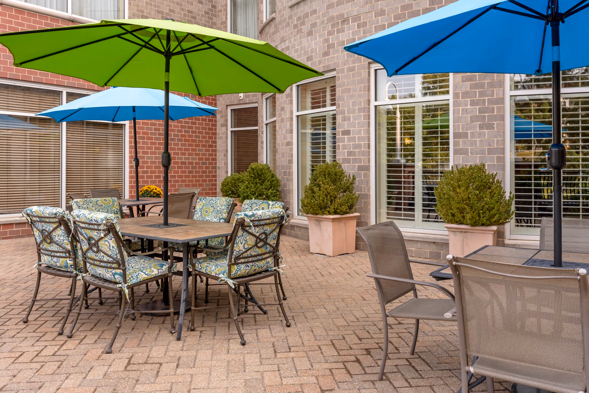 Patio courtyard with tables and patterned chairs under green and blue umbrellas beside a brick building.