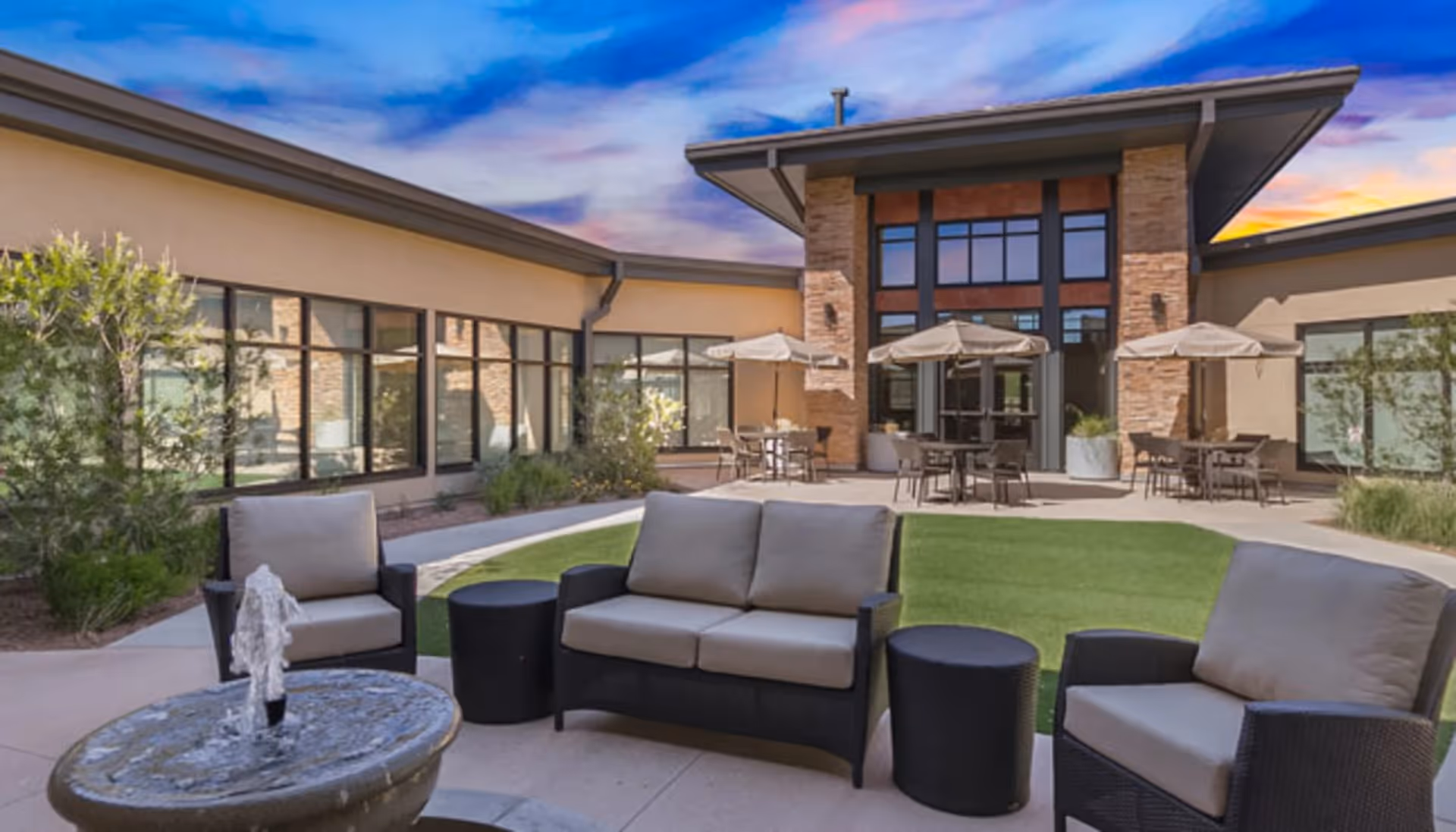 Outdoor courtyard area with cushioned patio furniture including a sofa and chairs arranged around a small water fountain. There are tables with umbrellas near the building, which has large windows and stone accents under a vibrant blue and purple sky.