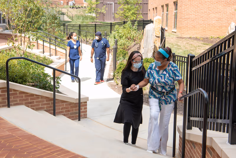 An outdoor walkway at a senior living facility with two women wearing face masks walking up a ramp, one assisting the other. In the background, two healthcare workers in scrubs and masks walk along a path surrounded by greenery and brick buildings.
