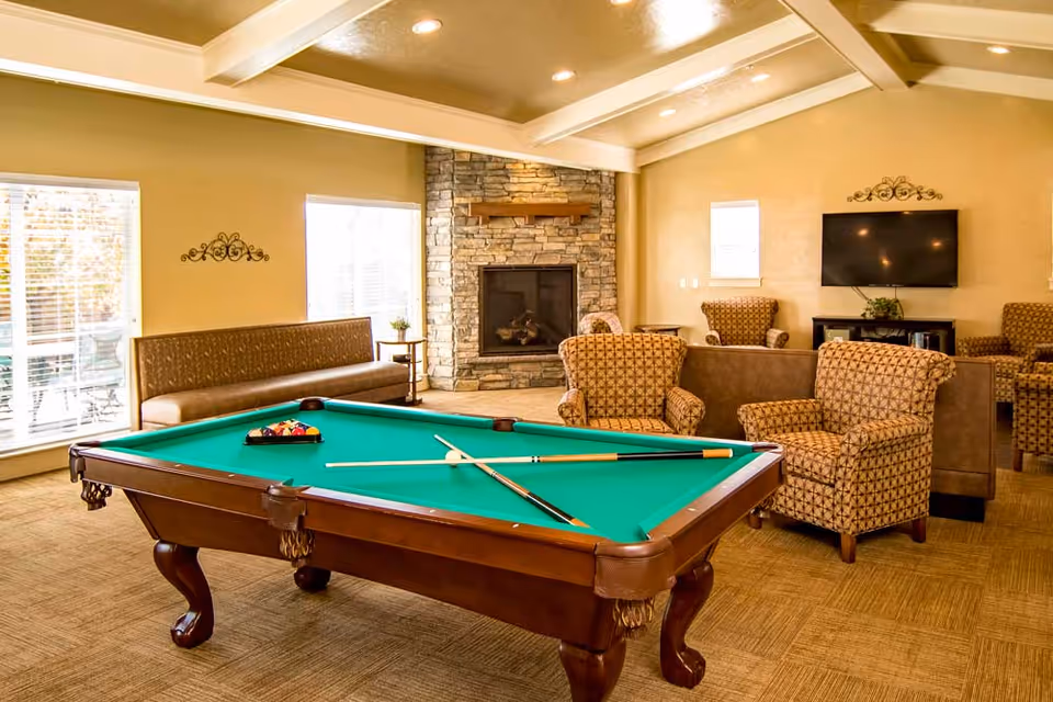 Bright common room with a pool table in the foreground, upholstered armchairs, a stone fireplace and a wall-mounted TV.