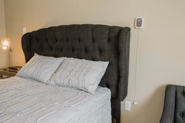A bedroom with a dark tufted headboard, a bed made with white textured bedding and two pillows, a bedside table with a lamp and books, and a wall-mounted emergency call button with a pull cord.