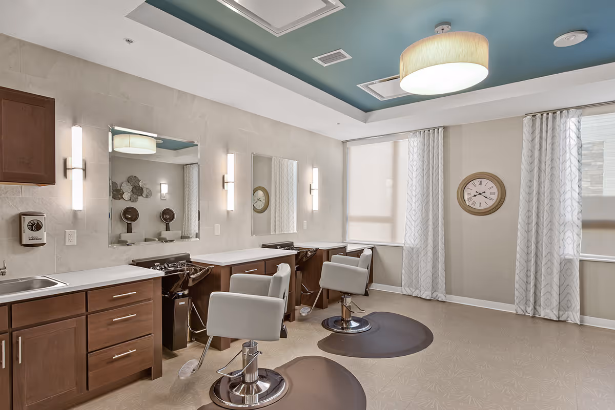 Interior view of a hair salon area in a senior living facility with two salon chairs in front of mirrors and sinks, wooden cabinets, a wall clock, and large windows with patterned curtains.
