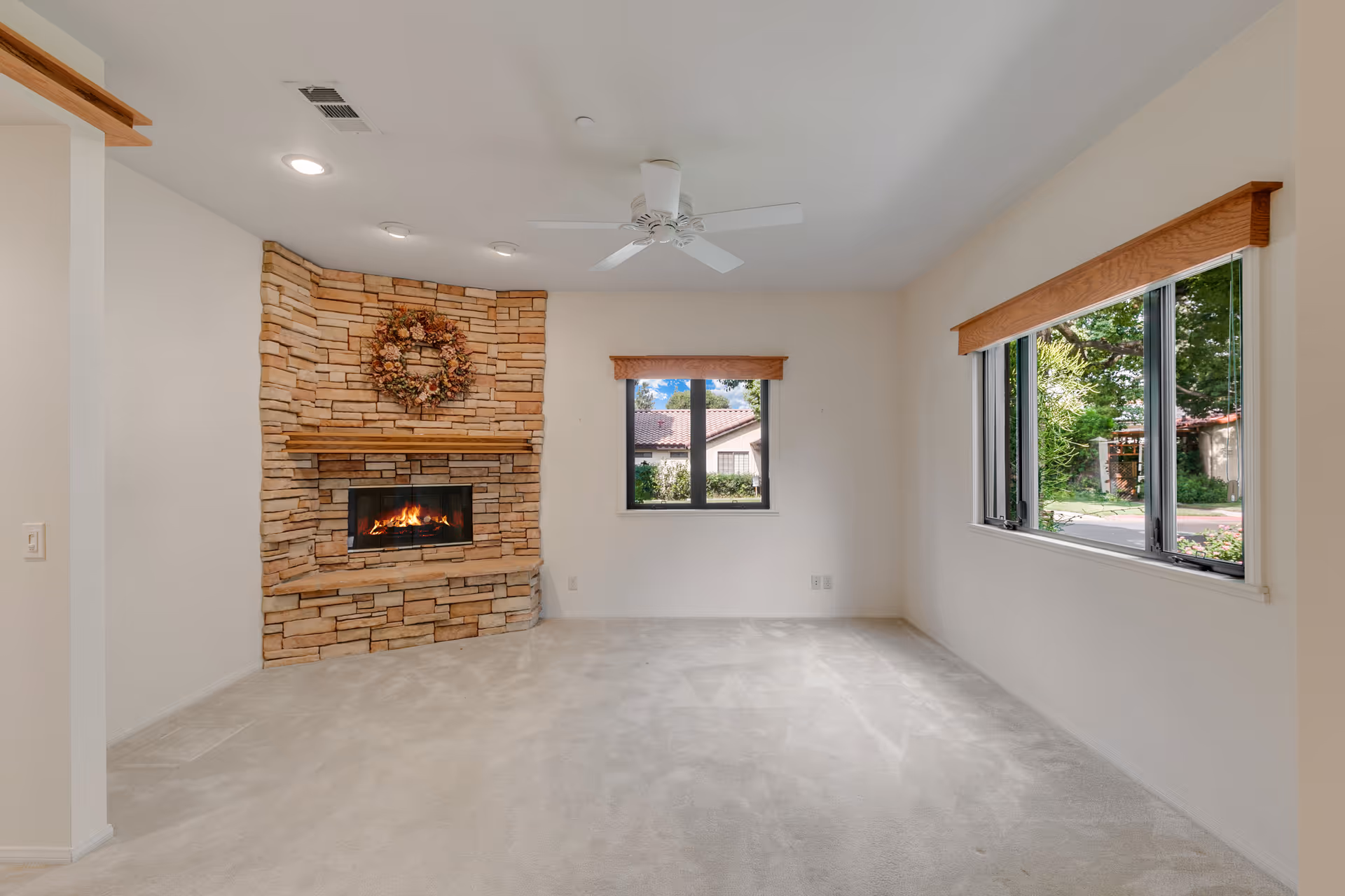 A bright living room with beige carpet, a stone fireplace with a wooden mantel and a floral wreath above it, a ceiling fan, and two windows with wooden valances showing greenery outside.
