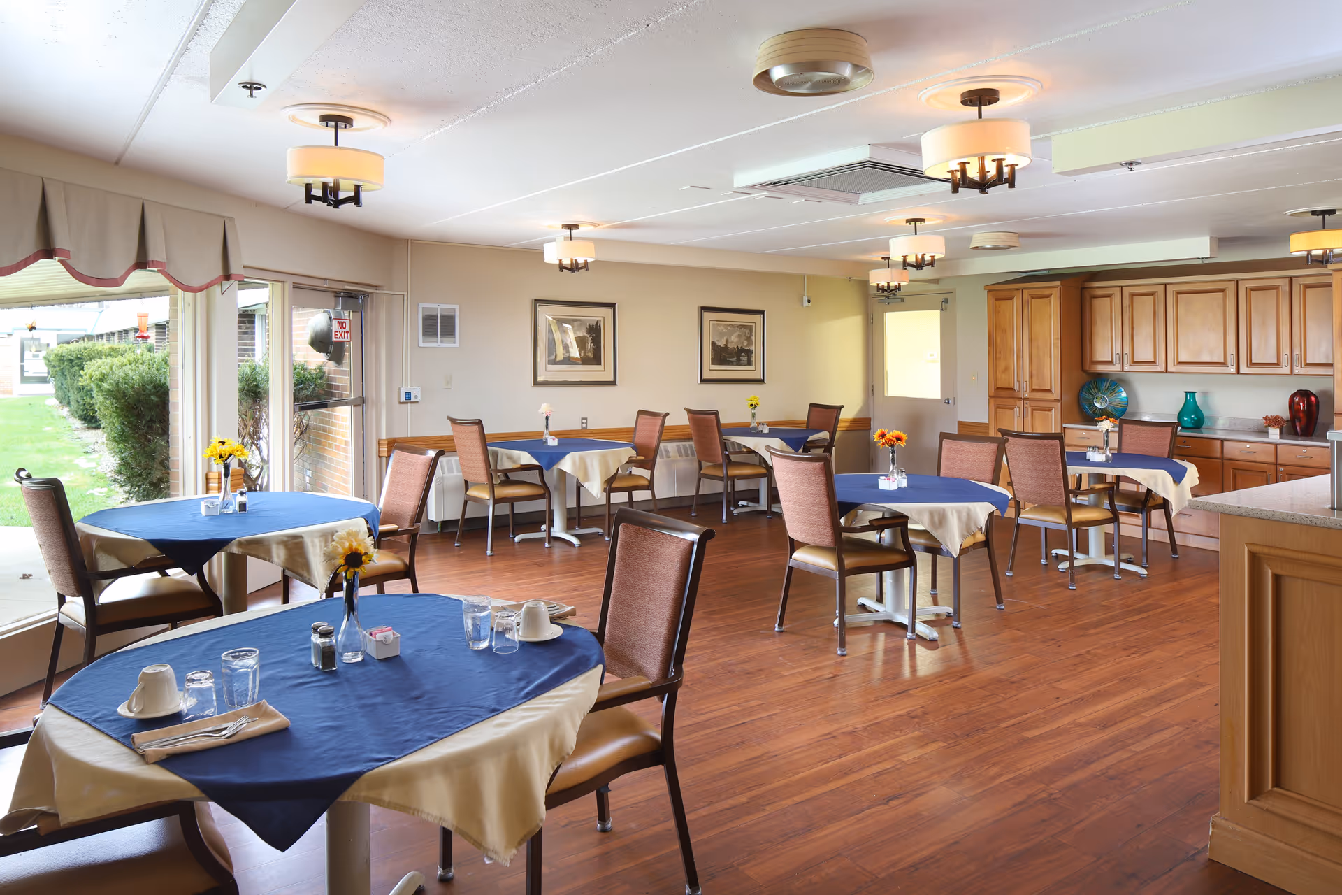 A bright dining room with several round tables covered with beige tablecloths and blue overlays, each set with glasses, cups, and small flower vases. The room has wooden flooring, beige walls with framed pictures, large windows with a view of greenery outside, and wooden cabinets along one wall. Ceiling lights provide warm illumination.