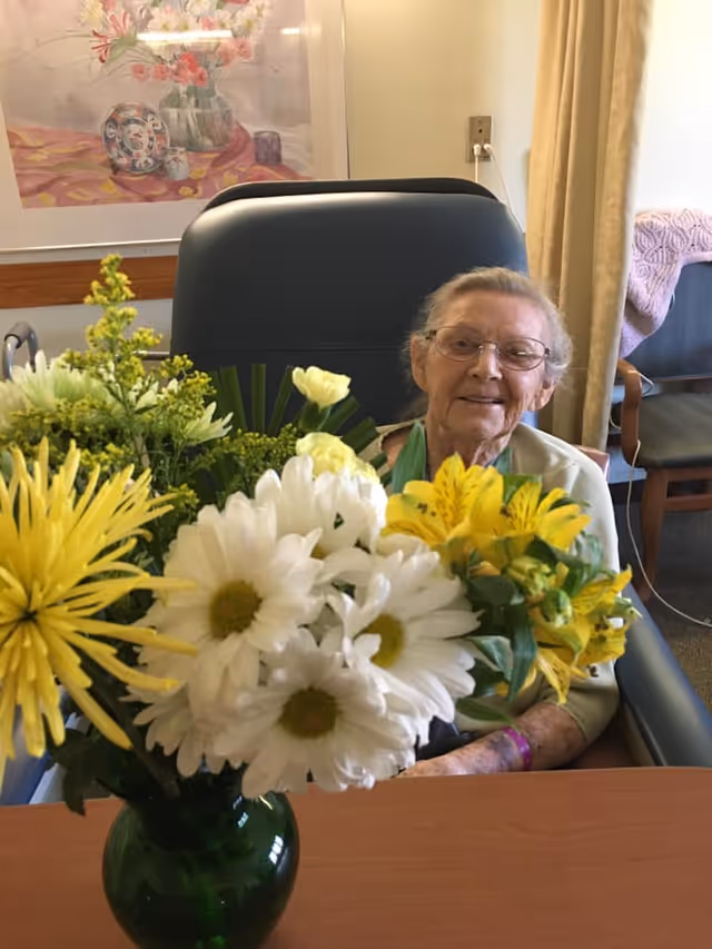 An elderly woman with glasses sitting in a chair behind a table with a green vase filled with yellow and white flowers. The room has a framed floral painting on the wall and a chair with a pink knitted blanket in the background.