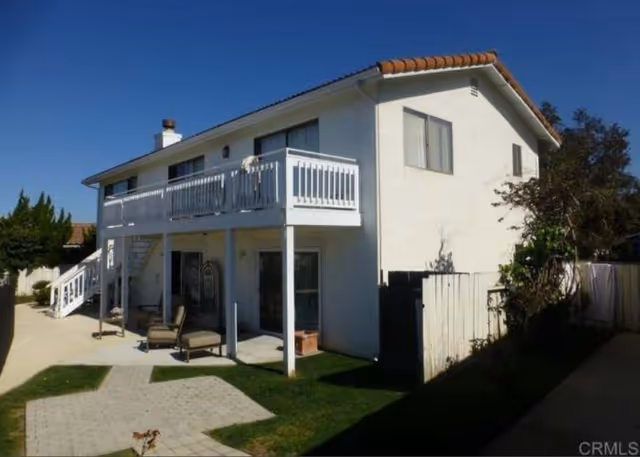 Two-story white residential building with a balcony on the upper floor and outdoor seating area on the ground floor, surrounded by a small grassy yard and paved walkway under a clear blue sky.