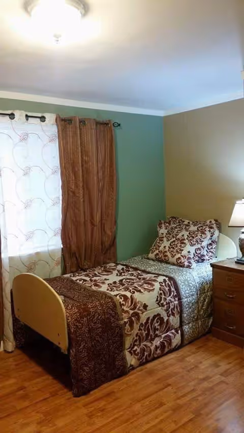 A small bedroom with a single bed covered in a patterned bedspread and matching pillows. The room has wooden flooring, a nightstand with a lamp, and two sets of curtains on the window, one sheer white with a circular pattern and one solid brown.