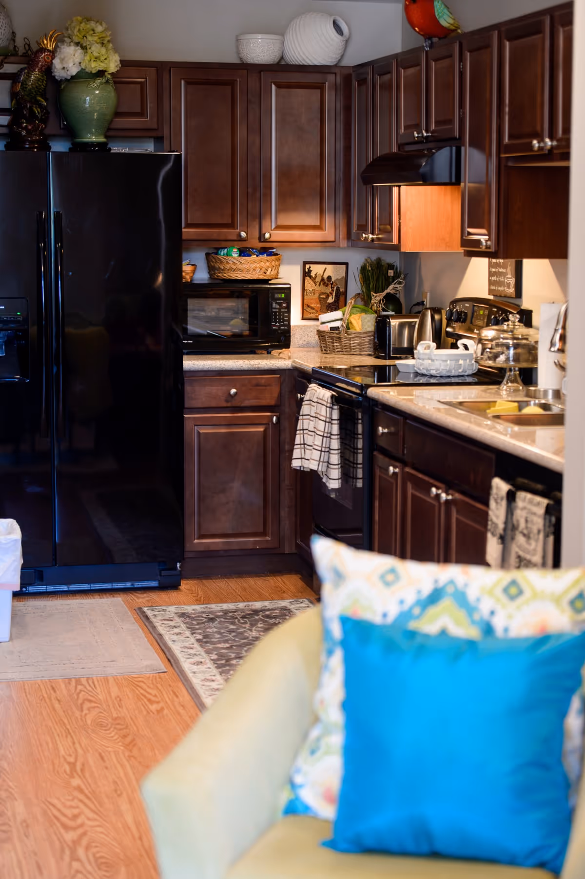 Cozy kitchen with dark wood cabinets, a black refrigerator and stove, countertop appliances, and a colorful pillowed chair in the foreground.