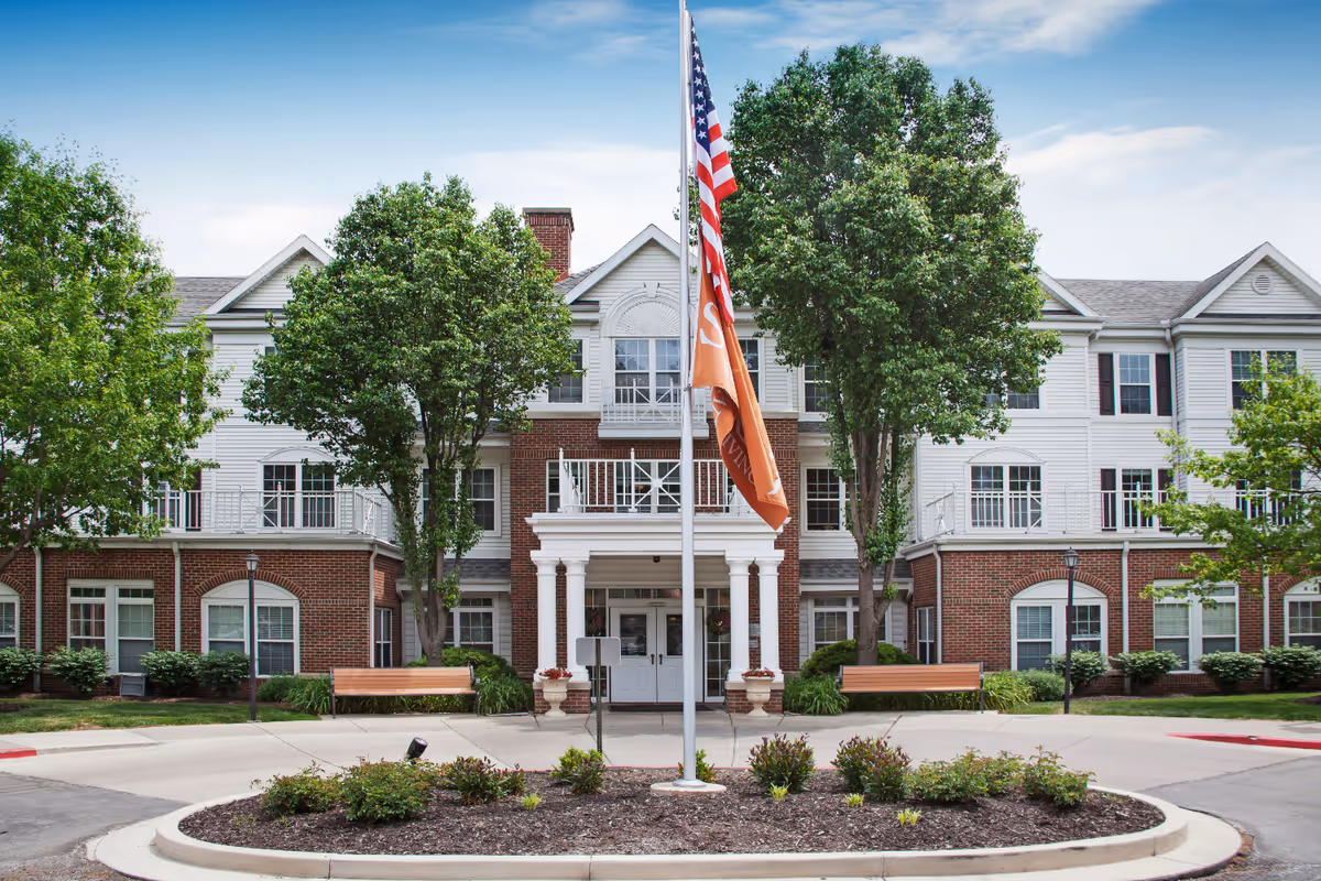 Entrance to a three-story brick-and-siding senior living building with flags, trees, benches, and a landscaped roundabout.