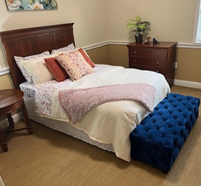 A neatly made bed with a wooden headboard in a bedroom. The bed has white sheets, floral pillowcases, and several decorative pillows in shades of red and pink. A pink throw blanket is draped across the foot of the bed. In front of the bed is a blue tufted bench. To the right of the bed is a wooden dresser with a potted plant and decorative items on top. A small wooden round side table is positioned to the left of the bed. The room has beige walls with white trim and a window on the right side.