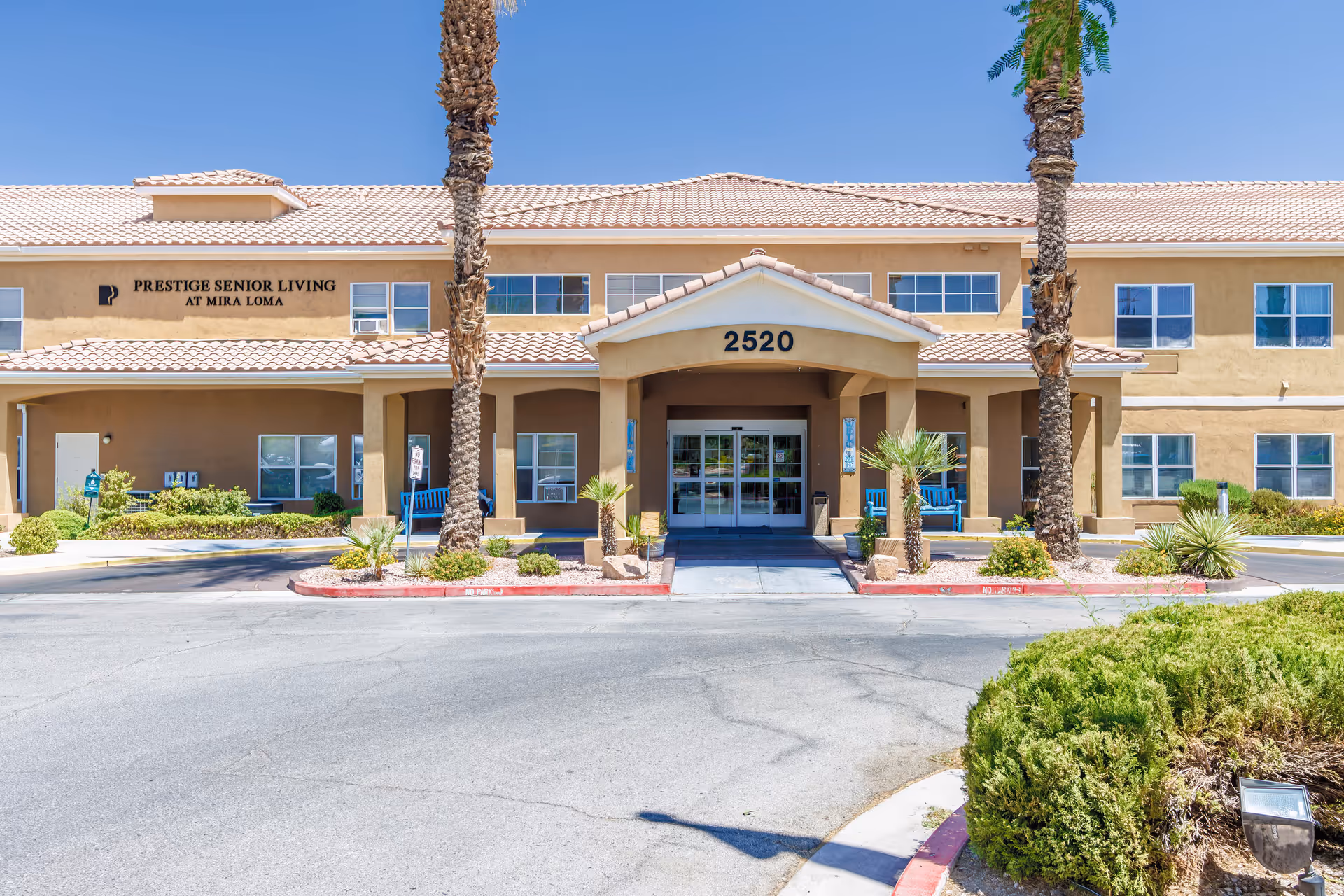 Front exterior view of a senior living facility building with beige walls and a tiled roof. The entrance has a covered porch with the number 2520 above it. There are palm trees and landscaped bushes around the driveway. The building has multiple windows and a sign that reads 'Prestige Senior Living at Mira Loma'.