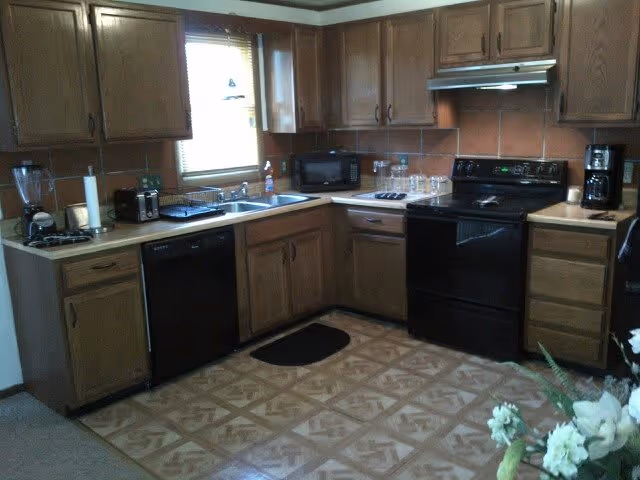 A kitchen with wooden cabinets, a black dishwasher, black stove with oven, microwave, coffee maker, blender, and a double sink under a window with blinds. The floor has a patterned linoleum covering and there is a small black mat in front of the sink. A vase with white flowers is partially visible in the foreground.