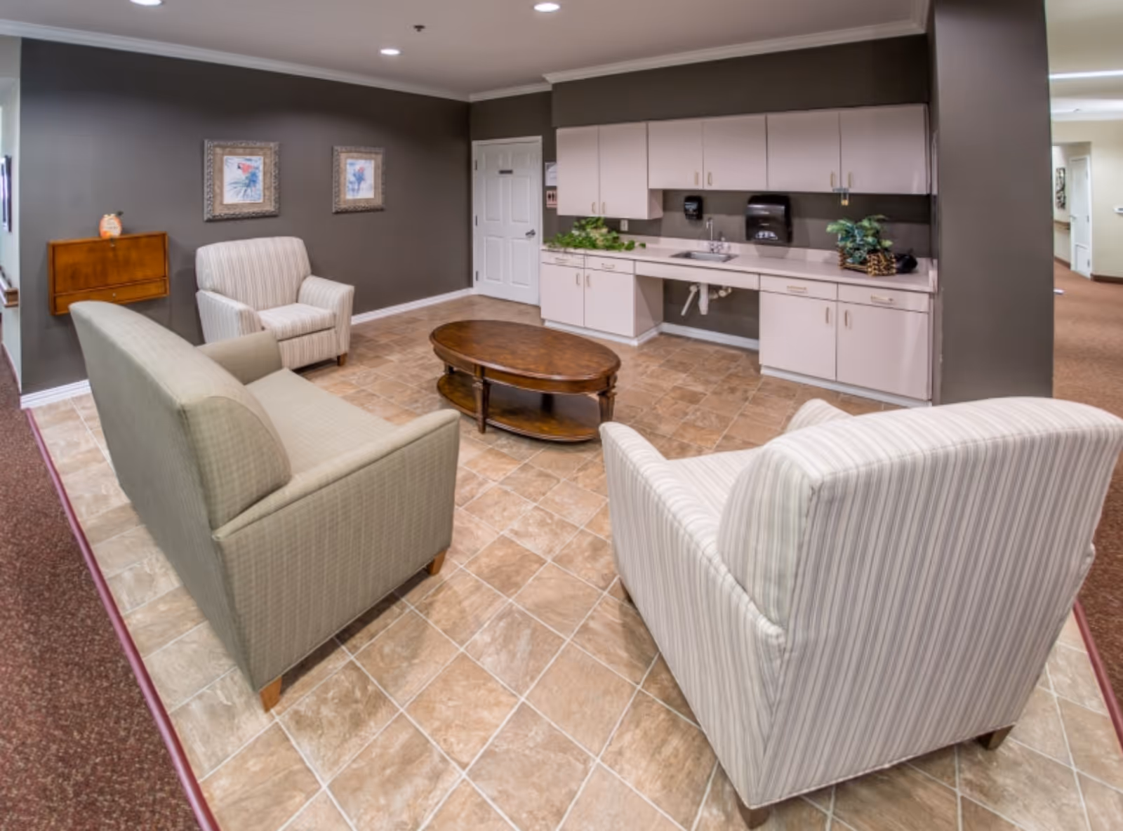 A cozy communal lounge with upholstered chairs and a sofa arranged around a wooden coffee table and a kitchenette along the back wall.