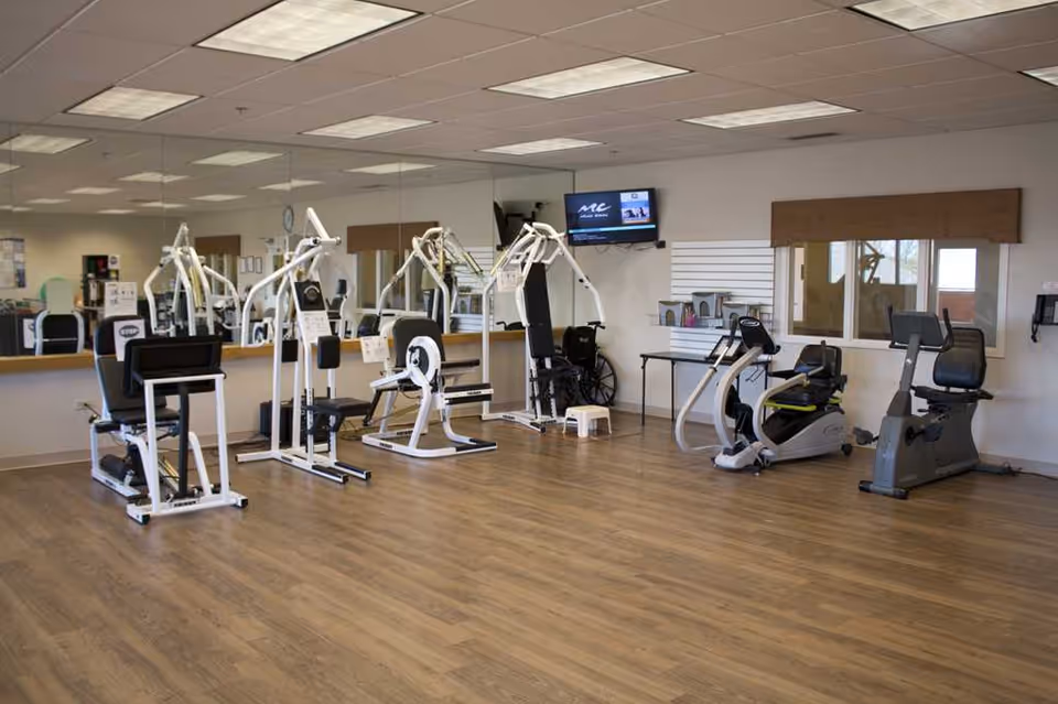 A fitness room with exercise equipment including stationary bikes, weight machines, and a wheelchair. The room has wooden flooring, a large mirror on one wall, a wall-mounted TV, and windows with brown valances.