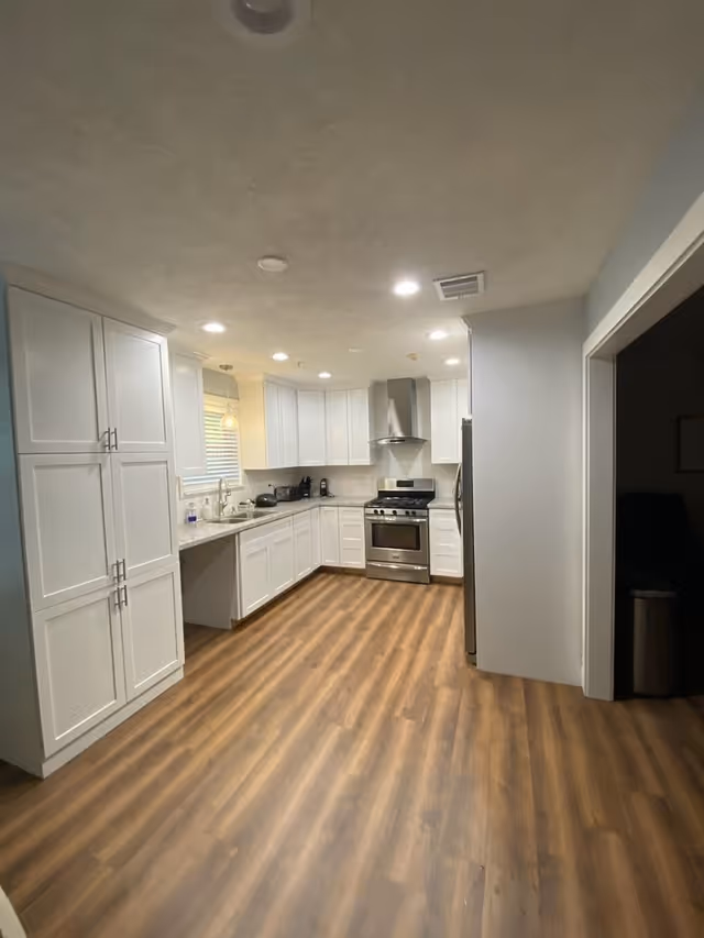 A modern kitchen with white cabinets, stainless steel appliances including a stove and refrigerator, a sink under a window, and wood-patterned flooring. The kitchen is well-lit with recessed ceiling lights and a pendant light above the sink.